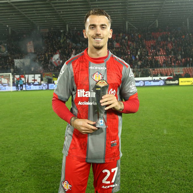 A male soccer player with short dark hair stands on a green grass field in a stadium under artificial lights, wearing a red and gray long-sleeve uniform with number 27 on the back, sponsor logos including Acerbi and Itas, holding a tall silver trophy with a base in both hands, smiling at the camera, with red stadium seats, advertising boards, and light smoke visible in the background.