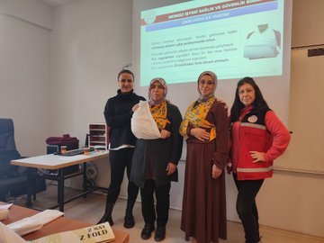 First image shows a group of women in headscarves and modest clothing seated in a classroom with filing cabinets and chairs holding a baby-sized first aid mannequin during training while a man in a red emergency vest demonstrates. Second image displays four women including some in headscarves and one in a red emergency vest standing in front of a projector screen with Turkish text about first aid training for women and family one holding a cloth bundle. Third image depicts a man and woman in red emergency vests demonstrating a head-tilt chin-lift maneuver on a mannequin to a seated group of women in headscarves in a room with tables and decorations.