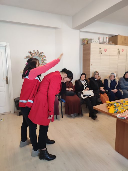 First image shows a group of women in headscarves and modest clothing seated in a classroom with filing cabinets and chairs holding a baby-sized first aid mannequin during training while a man in a red emergency vest demonstrates. Second image displays four women including some in headscarves and one in a red emergency vest standing in front of a projector screen with Turkish text about first aid training for women and family one holding a cloth bundle. Third image depicts a man and woman in red emergency vests demonstrating a head-tilt chin-lift maneuver on a mannequin to a seated group of women in headscarves in a room with tables and decorations.