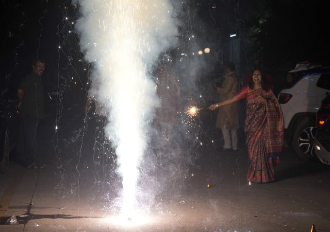 A nighttime scene outside with several people gathered near a white SUV car. A woman in a red and gold saree holds a lit sparkler emitting bright white sparks upward. Another man in traditional attire stands nearby watching. Smoke trails rise from the sparkler against a dark background with blurred lights in the distance.