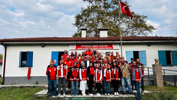 First image shows various blue toolboxes and bags from Makita brand arranged on a concrete floor in an indoor setting with shelves and banners in the background, including stacked cardboard boxes and a person handling items. Second image depicts a large group of people in red uniforms and casual attire posing together outdoors in front of a single-story building with blue shutters, red banner reading Düzce MEM AKUB, Turkish flag on a pole, and green grassy area.