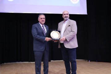 First image shows a man in suit speaking at podium with microphone in conference hall screen displays Turkiye Yuzyili Maarif Modeli and Hayat Boyu Ogrenme logos banners with Bingol University emblem and Turkish flags visible. Second image depicts large group of men and women in formal attire seated in auditorium rows some wearing headscarves table with white flowers and water glasses in front. Third image features diverse group of young women and men standing on stage in casual and formal clothes behind screen showing Ale Konferanslari Bingol logo. Fourth image portrays two men in suits one presenting plaque to the other on wooden floor against dark background with screen showing emblem.