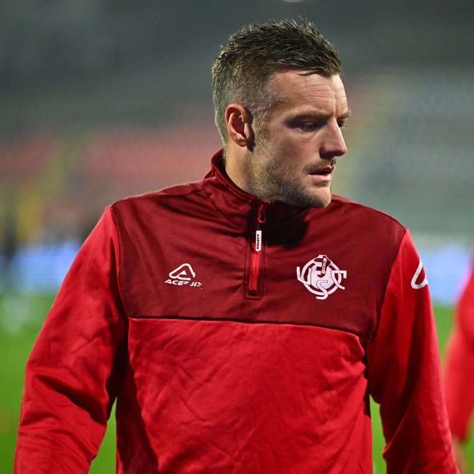 Man with short brown hair wearing red zip-up jacket with white accents and team crest on chest stands on green soccer field at night under stadium lights, blurred background of field and stands.