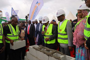 Group of officials including men in suits and safety vests with hard hats and women in hijabs and traditional dresses stand together at an outdoor event near construction site with banners reading MANCON and green tents in background. Several officials in yellow safety vests and hard hats lay hands on a stack of fresh concrete blocks during a groundbreaking ceremony with construction equipment and flags visible. Officials including men in suits and vests with hard hats gather around concrete blocks with one speaking into a microphone near tents and banners at the construction site. Group of officials in suits and traditional attire shake hands under white tents with green decorations at an outdoor ceremony.