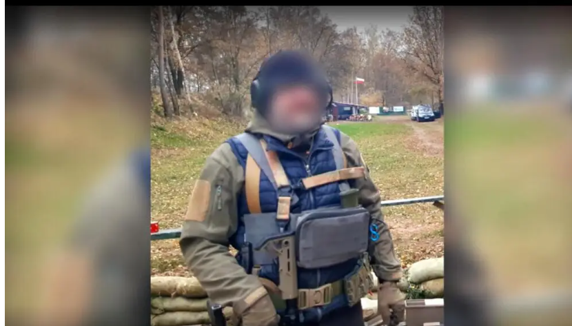 Person in tactical vest with shoulder straps, blue shirt, gray pouch on chest, tan arm patches, gray pants, brown gloves, black balaclava covering face, standing in forested area with trees, vehicles in background, red and white barrier, grass and dirt ground.