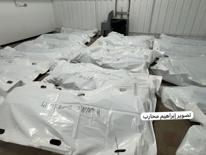 Multiple white body bags laid out on a concrete floor in an indoor facility with Arabic labels attached showing names dates and numbers like محمد أحمد النجار 17/10/2023 م and ح م م د ع ب د ا ل ر ح م ن الغول 17/10/2023 م some bags partially unzipped revealing shrouded forms stacked near walls and bins in a storage area.