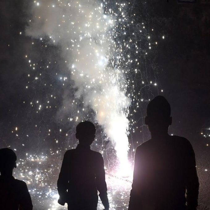 Silhouettes of several people standing and watching bursts of fireworks exploding in the night sky with smoke trails visible against a dark background the figures include adults and possibly children in casual clothing positioned on the ground near the fireworks launch area