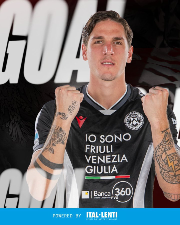 Nicolo Zaniolo stands celebrating with arms raised and fists clenched wearing a black Udinese jersey with white accents and sponsor logos including Banche 360 and Io Sono Friuli Venezia Giulia. Tattoos are visible on his arms and neck. Large white text GOAL appears behind him in the background. The jersey features the Udinese club emblem on the left chest.
