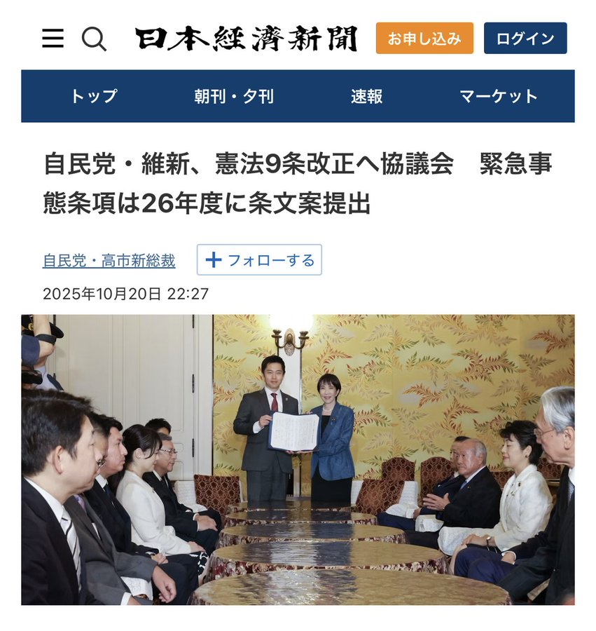 Image shows a group of Japanese politicians in a formal meeting room with yellow patterned walls and lamps. Several men and women in suits are seated around a large table, some holding documents. A central man in a suit stands presenting a document to a woman in blue attire. The scene appears official with participants including older men and women engaged in discussion.