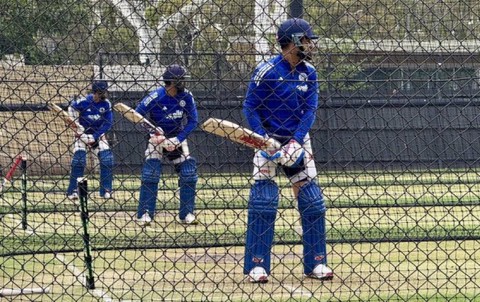 Three Indian cricketers, Virat Kohli, Rohit Sharma, and Shubman Gill, stand on a grassy practice field enclosed by a black fence and nets. They wear blue team uniforms, long-sleeve shirts, pants, and leg pads, holding wooden cricket bats. Kohli is in the foreground facing forward with a helmet, Sharma and Gill slightly behind preparing to bat. Red cones mark the boundary.