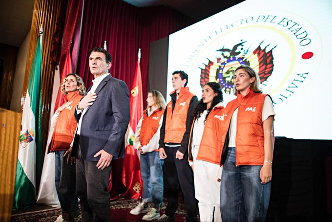 A group of people stands on a stage with red curtains and wooden columns behind a large screen displaying the text Eleccion del Estado and the Bolivian coat of arms several Bolivian flags are positioned around the stage individuals wear orange vests and formal attire including suits and casual clothing one man in a dark suit stands prominently with hand on chest others pose respectfully in a formal gathering setting