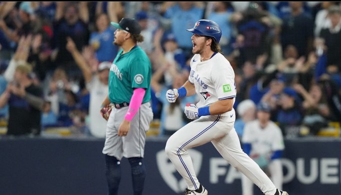 Two baseball players in uniforms celebrate a victory one in Seattle Mariners green jersey with arm extended and glove on hand the other in Toronto Blue Jays white jersey with blue accents raising arms and glove while wearing batting helmet surrounded by blurred stadium crowd and field in background.