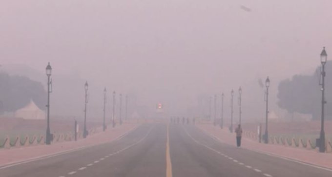 A wide straight road stretches through a hazy urban area with reduced visibility due to thick grey fog covering the scene. Tall ornate street lamps line both sides of the road spaced evenly amid sparse trees. A few distant figures including people and vehicles are barely visible in the mist ahead. The atmosphere appears polluted with low light suggesting early morning or poor air quality.