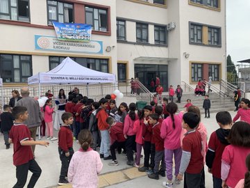 First image shows a group of adults and children standing together under a white tent labeled Manisa İl Milli Eğitim Müdürlüğü outside a school building with yellow walls and a sign for Vicdan Karaosmanoğlu school children wearing red uniforms holding balloons and standing around a table with papers. Second image depicts teenage girls in red uniforms holding blue signs that say something about health under another white tent with tables covered in colorful game boards and stacks of brochures near a school railing. Third image features a large group of children mostly in pink and red clothing gathered around a white tent outside the school entrance with a blue banner and trash bin visible in the background. Fourth image displays children in school uniforms including red polo shirts interacting with a large pink tooth model and toothbrushes on a white table covered with health logos and stacks of books under the tent.