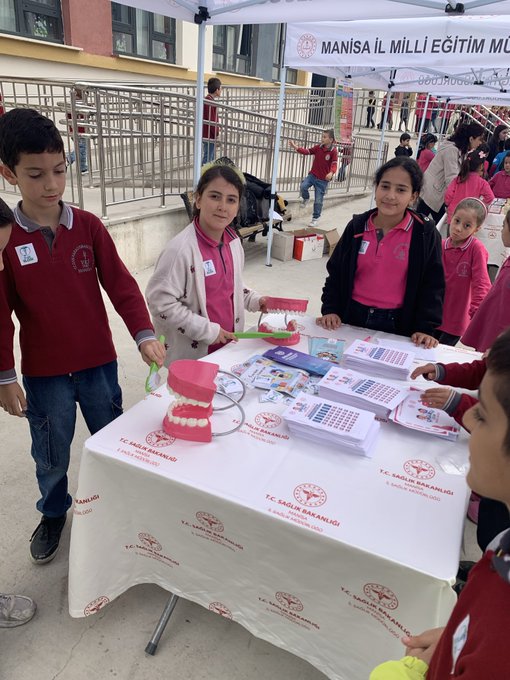 First image shows a group of adults and children standing together under a white tent labeled Manisa İl Milli Eğitim Müdürlüğü outside a school building with yellow walls and a sign for Vicdan Karaosmanoğlu school children wearing red uniforms holding balloons and standing around a table with papers. Second image depicts teenage girls in red uniforms holding blue signs that say something about health under another white tent with tables covered in colorful game boards and stacks of brochures near a school railing. Third image features a large group of children mostly in pink and red clothing gathered around a white tent outside the school entrance with a blue banner and trash bin visible in the background. Fourth image displays children in school uniforms including red polo shirts interacting with a large pink tooth model and toothbrushes on a white table covered with health logos and stacks of books under the tent.