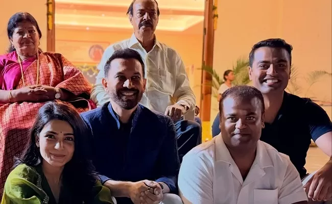 Group of six people posing indoors near open doors with warm lighting and plants in background. Two elderly women in red and green sarees with jewelry sit on left. Central man in white kurta sits smiling. Woman in pink top and black hair sits beside man in dark shirt. Two younger men in white and black shirts stand smiling on right.