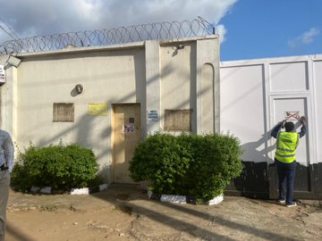 First image shows a beige building with barbed wire fencing under a partly cloudy sky, featuring a yellow sign reading 360, small windows, a brown door, green bushes, and two workers in hi-vis vests, one sealing with an X mark. Second image depicts a worker in hi-vis vest painting an X mark on a white wall next to a door on a similar fenced structure with green bushes and ground. Third image displays several workers in hi-vis vests outside a yellow building with barred door, handling white spherical objects in black buckets and wooden stands amid various tools and containers. Fourth image illustrates a worker in hi-vis vest posting X-marked notices on a barred metal door of a yellow building, with additional notices and a bucket of white substance nearby.