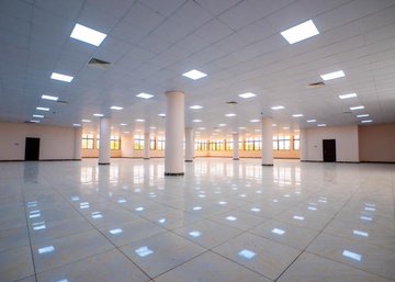 Aerial view of a large four-storey university library building with beige walls, red roofs, decorative patterns, a prominent green dome on top, surrounded by parking lots with vehicles, people walking, green lawns, and adjacent red-roofed structures in a urban campus setting. Interior shot of a spacious computer laboratory with multiple rows of green desks, purple partitions, computer monitors on tables, overhead fluorescent lighting, beige columns, and air conditioning units. Multi-level atrium inside the library featuring a circular design with white walls, glass railings on upper floors, a central open space with blue-green skylight dome, escalators or stairs, and potted plants. Expansive empty hall with shiny green tiled floors, white walls, large windows allowing natural light, beige columns, doors on sides, and ceiling lights.
