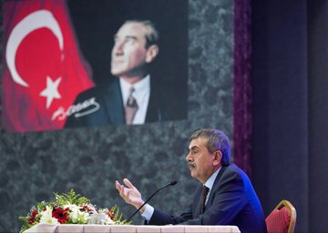 First image shows a man in a suit speaking at a podium with a microphone hand raised gesturing toward a large screen displaying a portrait of Recep Tayyip Erdoğan with Turkish flags on stage surrounded by flowers and a formal table setting. Second image depicts the same man in a suit standing at a podium addressing a large audience of suited individuals seated in rows in a lit auditorium with Turkish flags and banners on walls. Third image features a stage with banners reading İlçe Millî Eğitim Müdürleri Toplantısı showing the man seated at a table with flowers and a screen displaying Erdoğan portrait and Turkish flags with audience seated in chairs. Fourth image captures a large group of formally dressed men and women standing in rows in a hall with flowers on a table and stage lighting overhead.