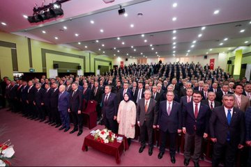 First image shows a man in a suit speaking at a podium with a microphone hand raised gesturing toward a large screen displaying a portrait of Recep Tayyip Erdoğan with Turkish flags on stage surrounded by flowers and a formal table setting. Second image depicts the same man in a suit standing at a podium addressing a large audience of suited individuals seated in rows in a lit auditorium with Turkish flags and banners on walls. Third image features a stage with banners reading İlçe Millî Eğitim Müdürleri Toplantısı showing the man seated at a table with flowers and a screen displaying Erdoğan portrait and Turkish flags with audience seated in chairs. Fourth image captures a large group of formally dressed men and women standing in rows in a hall with flowers on a table and stage lighting overhead.