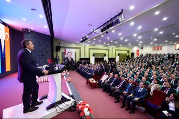 First image shows a man in a suit speaking at a podium with a microphone hand raised gesturing toward a large screen displaying a portrait of Recep Tayyip Erdoğan with Turkish flags on stage surrounded by flowers and a formal table setting. Second image depicts the same man in a suit standing at a podium addressing a large audience of suited individuals seated in rows in a lit auditorium with Turkish flags and banners on walls. Third image features a stage with banners reading İlçe Millî Eğitim Müdürleri Toplantısı showing the man seated at a table with flowers and a screen displaying Erdoğan portrait and Turkish flags with audience seated in chairs. Fourth image captures a large group of formally dressed men and women standing in rows in a hall with flowers on a table and stage lighting overhead.