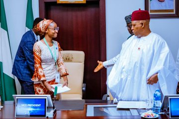 First image shows a woman in orange headwrap and patterned dress standing near a desk with Nigerian flags and laptops labeled President and Vice President, engaging with a man in white traditional attire and red cap extending his hand in greeting, in a formal office setting. Second image depicts the same woman seated at a desk with laptops and water bottles, discussing with the man in white attire and red cap writing notes, surrounded by Nigerian flags and official portraits. Third image features a group of women and girls in hijabs and traditional dresses with lanyards posing with the man in white attire and red cap on stairs in a building with plants. Fourth image captures a large diverse group of men and women in various traditional and formal outfits including blue suits, colorful dresses, and white attire, standing together in a spacious lobby with palm trees and modern architecture.