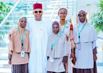First image shows a woman in orange headwrap and patterned dress standing near a desk with Nigerian flags and laptops labeled President and Vice President, engaging with a man in white traditional attire and red cap extending his hand in greeting, in a formal office setting. Second image depicts the same woman seated at a desk with laptops and water bottles, discussing with the man in white attire and red cap writing notes, surrounded by Nigerian flags and official portraits. Third image features a group of women and girls in hijabs and traditional dresses with lanyards posing with the man in white attire and red cap on stairs in a building with plants. Fourth image captures a large diverse group of men and women in various traditional and formal outfits including blue suits, colorful dresses, and white attire, standing together in a spacious lobby with palm trees and modern architecture.