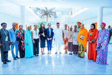 First image shows a woman in orange headwrap and patterned dress standing near a desk with Nigerian flags and laptops labeled President and Vice President, engaging with a man in white traditional attire and red cap extending his hand in greeting, in a formal office setting. Second image depicts the same woman seated at a desk with laptops and water bottles, discussing with the man in white attire and red cap writing notes, surrounded by Nigerian flags and official portraits. Third image features a group of women and girls in hijabs and traditional dresses with lanyards posing with the man in white attire and red cap on stairs in a building with plants. Fourth image captures a large diverse group of men and women in various traditional and formal outfits including blue suits, colorful dresses, and white attire, standing together in a spacious lobby with palm trees and modern architecture.
