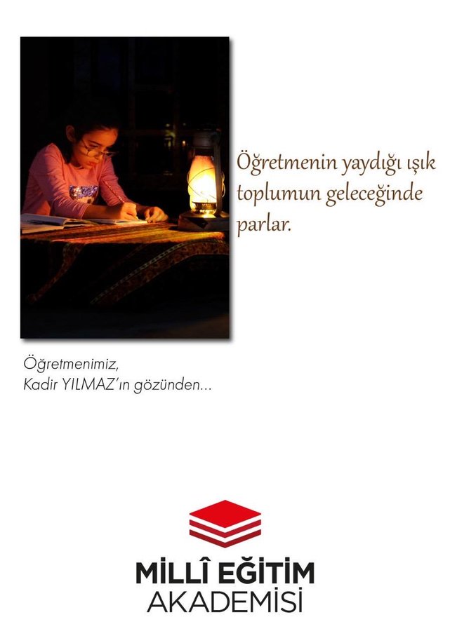 A woman in pink clothing sits at a wooden desk with books and a notebook, writing under the warm glow of a lit lantern placed on the desk. She appears focused and dedicated in a dimly lit room setting. Turkish text overlays the image stating that the light kindled by the teacher illuminates society and the future, with additional phrasing about teachers from our chain. The Milli Egitim Akademisi logo in red is at the bottom right.