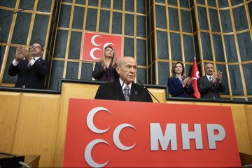 First image shows eight formally dressed individuals including men in suits and one woman in a dark outfit standing on a paved area with large stone columns and a building entrance in the background under clear skies. Second image depicts a large wooden-paneled hall filled with numerous people mostly men in dark suits seated and standing with some holding cameras and microphones clustered around a central figure at a podium illuminated by chandeliers. Third image features a close-up of an elderly man with bald head and glasses wearing a dark suit white shirt and patterned tie looking serious against a blurred background. Fourth image displays several people including suited men and women raising hands in applause on a balcony with Turkish flags and a large red banner reading MHP behind a wooden podium in a ornate hall with wooden paneling.