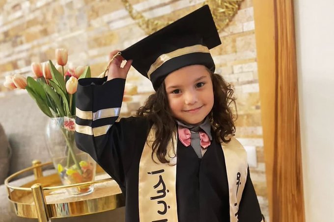 A young girl with curly brown hair smiles while wearing a black graduation cap and gown with gold trim over a white shirt and bow tie standing in a room with beige brick walls and a gold side table holding a vase of pink tulips she holds the cap in one hand and raises the other arm a sash with Hebrew text drapes across her chest.