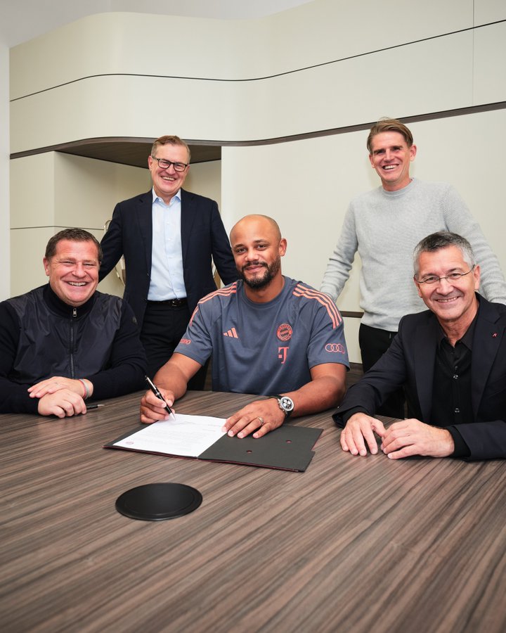 Five men in a modern conference room with curved walls and wooden table. Leftmost man in dark jacket and blue shirt smiling. Next man bald with goatee in gray Bayern Munich jersey signing document with pen. Central man in gray sweater smiling. Right two men in suits one smiling one serious. Documents and black circular object on table.