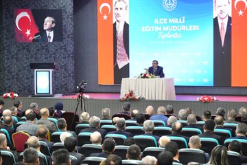 First image displays a conference stage with large screens showing Turkish flags and text İLÇE MİLLİ EĞİTİM MÜDÜRLERİ TOPLANTISI backdrop featuring portraits of suited men one speaking at podium with microphone flowers on table audience of men in suits seated in rows chairs and projector screen visible. Second image shows wide view of auditorium with green walls lighting rigs large portrait of suited man with Turkish flag audience of men and women in formal attire seated in rows speaker at table with microphone flowers and documents on stage. Third image focuses on man in dark suit with red lanyard holding microphone speaking to audience of men in suits seated in green chairs backdrop with blurred stage elements. Fourth image depicts man in suit with lanyard holding microphone speaking audience of men in suits seated behind him green wall with red emblem and Arabic script visible.