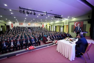 First image displays a conference stage with large screens showing Turkish flags and text İLÇE MİLLİ EĞİTİM MÜDÜRLERİ TOPLANTISI backdrop featuring portraits of suited men one speaking at podium with microphone flowers on table audience of men in suits seated in rows chairs and projector screen visible. Second image shows wide view of auditorium with green walls lighting rigs large portrait of suited man with Turkish flag audience of men and women in formal attire seated in rows speaker at table with microphone flowers and documents on stage. Third image focuses on man in dark suit with red lanyard holding microphone speaking to audience of men in suits seated in green chairs backdrop with blurred stage elements. Fourth image depicts man in suit with lanyard holding microphone speaking audience of men in suits seated behind him green wall with red emblem and Arabic script visible.