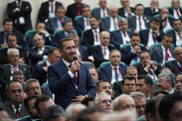 First image displays a conference stage with large screens showing Turkish flags and text İLÇE MİLLİ EĞİTİM MÜDÜRLERİ TOPLANTISI backdrop featuring portraits of suited men one speaking at podium with microphone flowers on table audience of men in suits seated in rows chairs and projector screen visible. Second image shows wide view of auditorium with green walls lighting rigs large portrait of suited man with Turkish flag audience of men and women in formal attire seated in rows speaker at table with microphone flowers and documents on stage. Third image focuses on man in dark suit with red lanyard holding microphone speaking to audience of men in suits seated in green chairs backdrop with blurred stage elements. Fourth image depicts man in suit with lanyard holding microphone speaking audience of men in suits seated behind him green wall with red emblem and Arabic script visible.