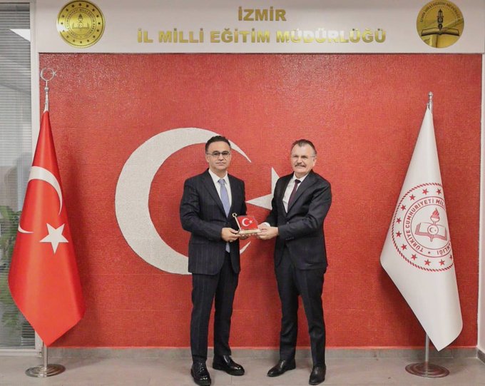 First image displays two men in dark suits shaking hands with plaques reading İl Milli Eğitim Müdürlüğü on a red wall flanked by Turkish flags and a white flag with a red emblem. Second image shows five men in suits including the two from the first image exchanging a document or plaque in the same formal setting with additional participants wearing ties and professional attire against the institutional backdrop with golden emblems.