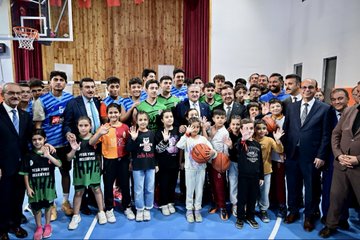 First image shows a large group of adults and children posing together in a gymnasium with wooden walls, red curtains, and basketball hoops; many wear green or blue sports jerseys, some hold basketballs, all smiling and standing on a blue court. Second image depicts two suited men, one holding a wrapped red and gold embroidered textile gift with orange ribbon, standing before a Turkish flag and a gold sign reading AK PARTI IL BASKANLIGI, with another man in suit nearby. Third image captures a group of formally dressed men and women in suits and traditional attire standing on steps in front of a beige building labeled MALATYA VALİLİĞİ with Turkish flag elements. Fourth image features a line of men in dark suits standing on blue carpeted steps outside a building with a white circle logo marked 100 YIL, modern architecture in background.