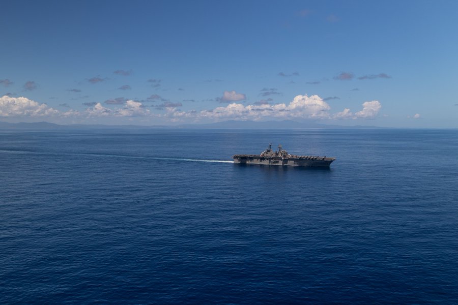 A large amphibious assault ship identified as USS Iwo Jima navigates through open ocean waters under a clear blue sky with scattered white clouds. The vessel features a flat deck with aircraft visible and a prominent island superstructure. It leaves a white wake trailing behind as it moves forward in the Caribbean Sea during a military exercise.