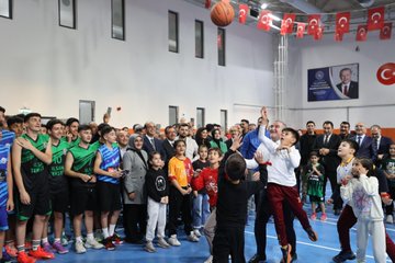 First image shows a man in a suit speaking at a podium during the opening ceremony with a large screen displaying Turkish text Yakınca Gençlik Merkezi and Tenis Kortu Açılış Töreni in Malatya along with microphones and banners. Second image depicts the same man in a suit holding a ribbon with children including boys and girls some wearing headscarves and casual clothes surrounded by adults in formal attire cutting the ribbon in front of a stage. Third image captures a group of young basketball players in green jerseys and others in casual clothes with adults including suited officials jumping to shoot a basketball into a hoop inside a gymnasium with Turkish flags and banners on the walls. Fourth image features a large group photo of children in sports jerseys holding basketballs adults in suits and formal wear posing together in a hall with wooden paneling red curtains a basketball hoop and Turkish flags in the background.