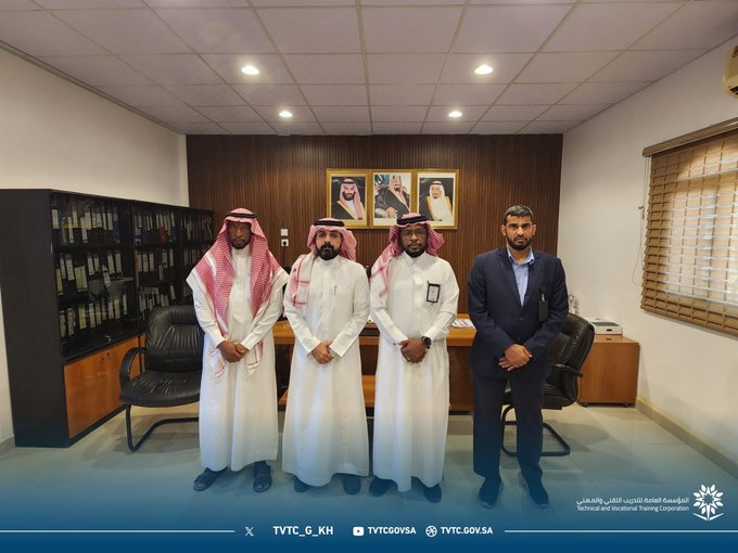 Four men in traditional white thobes and headscarves or suits stand in a modern office with wooden paneling, bookshelves, framed portraits on the wall, desks, chairs, and large windows. One man wears a blue suit and another has a badge on his thobe. The setting includes air conditioning units and office furniture.