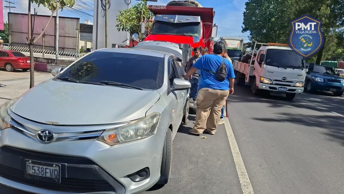 Silver Toyota sedan with license plate visible parked on roadside next to red dump truck in urban street setting with trees and other vehicles including white pickup truck and red car in background blue uniformed officer in blue shirt khaki pants and backpack stands at open driver door of sedan inspecting scene with PMT badge on vehicle and sign nearby under clear sky
