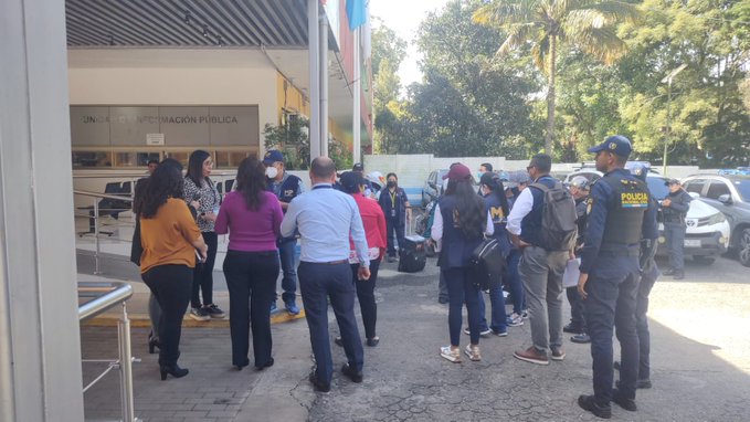 Group of people including officials in suits and police officers in vests with yellow markings stand and converse outside a modern building entrance with striped awning, palm trees, and parked vehicles in a sunny outdoor parking area.