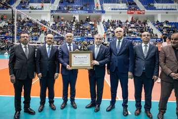 Six men in dark suits stand on indoor volleyball court holding framed certificate with gold emblem center man smiling others clapping orange court blue seating audience blurred background Turkish flags banners visible. Eight people including women in formal attire seated red chairs stands man in blue suit gesturing others watching attentively water bottle table foreground. Players in white blue uniforms compete volleyball match orange court referee in black players jumping spike net gym bright lights full stands spectators cheering. Large banner Malatya BGFB yellow black stripes crowd in yellow black clothing cheering steps holding phones cup trophies displayed area event signage.