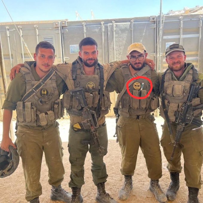 Four men in military uniforms and tactical vests stand together outdoors near shipping containers under a clear sky. They carry rifles slung over shoulders and wear boots and caps. A red circle highlights a badge on one soldiers chest. The group appears posed for a photo in a desert-like setting.