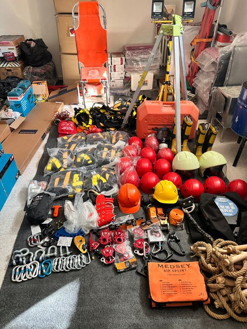 First image displays assortment of safety equipment on floor including multiple red helmets yellow hard hats orange backpacks black gloves red fire extinguishers orange stretcher yellow tripod stand ropes carabiners and storage boxes in indoor setting. Second image shows yellow stretcher on table alongside blue power tools orange chainsaw with Husqvarna branding black battery packs orange cable reel in protective case and white table surface. Third image features orange chainsaw yellow stretcher gray and black communication devices orange cable bag with DeWalt branding blue tool boxes on white table with person in background. Fourth image depicts blue floodlight on tripod yellow stretcher power tools storage boxes and person wearing black shirt and blue uniform near table in room with gray floor.