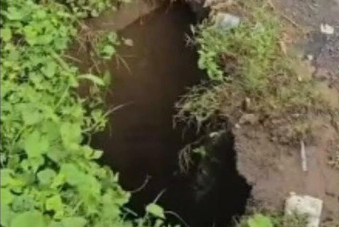 Una carretera pavimentada negra con un gran agujero rectangular en el centro causado por erosion del suelo debajo mostrando tierra expuesta y agua acumulada. Vegetacion verde como arbustos y hierbas crece abundantemente a lo largo de los bordes del agujero y en las orillas de la carretera. El terreno circundante es irregular con suelos expuestos y mas vegetacion. El puente Chantel esta daÃ±ado por las lluvias afectando la conexion entre El Quetzal y La Reforma en San Marcos.