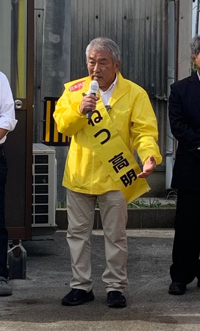 First image shows an elderly man with gray hair wearing a white shirt under a bright yellow raincoat-like vest labeled with black Japanese characters speaking into a microphone held in his right hand while gesturing with his left hand outdoors near a metal shutter door with other men in white shirts and suits standing nearby including one in a dark suit to his right and another in white to his left with yellow caution tape and an air conditioning unit visible in the background. Second image depicts two men one older with gray hair in a yellow vest and another younger in a dark suit both holding microphones standing in front of a small red car parked beside a building with a sign reading 〒754-1712 Yamaguchi Prefecture and a large yellow banner featuring the candidates face and Japanese text for election promotion with trees and a road in the background.
