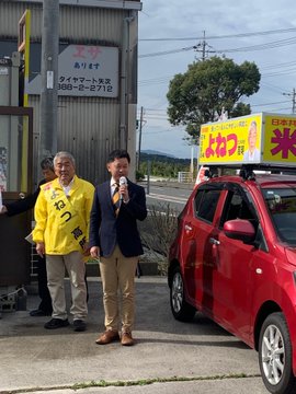 First image shows an elderly man with gray hair wearing a white shirt under a bright yellow raincoat-like vest labeled with black Japanese characters speaking into a microphone held in his right hand while gesturing with his left hand outdoors near a metal shutter door with other men in white shirts and suits standing nearby including one in a dark suit to his right and another in white to his left with yellow caution tape and an air conditioning unit visible in the background. Second image depicts two men one older with gray hair in a yellow vest and another younger in a dark suit both holding microphones standing in front of a small red car parked beside a building with a sign reading 〒754-1712 Yamaguchi Prefecture and a large yellow banner featuring the candidates face and Japanese text for election promotion with trees and a road in the background.