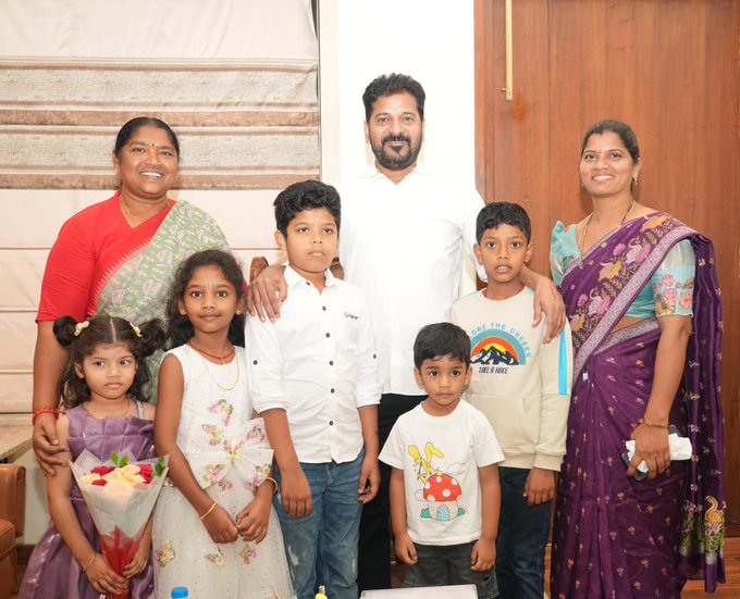 Group of eight people including four adults and four children stand together indoors against a beige curtain and wooden door. The adults wear traditional sarees and shirt, with one man in white. Children dressed in casual clothes like t-shirts and dresses, some holding flowers. All smiling at camera in a family pose.