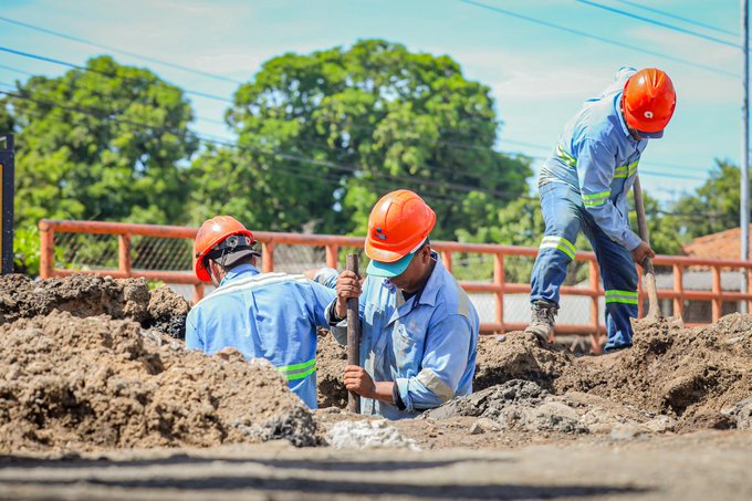 Three workers wearing blue uniforms with reflective strips and orange safety helmets engage in manual labor at a construction site. One worker holds a tool while digging into a pile of loose brown dirt. Another worker stands nearby assisting with the task. The background includes green trees, a metal fence, and clear blue sky.